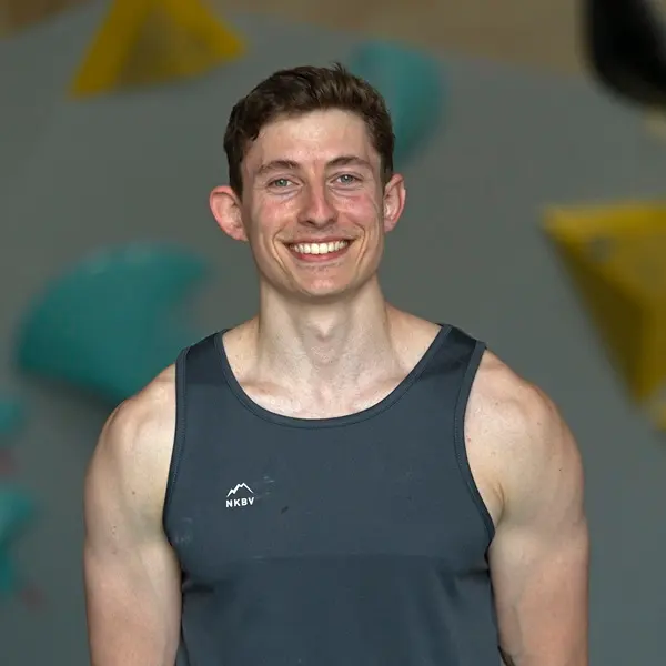 Een man met kort bruin haar en een grijze tanktop glimlacht naar de camera. Op de muur in de wazige achtergrond zijn felgekleurde klimgrepen te zien.